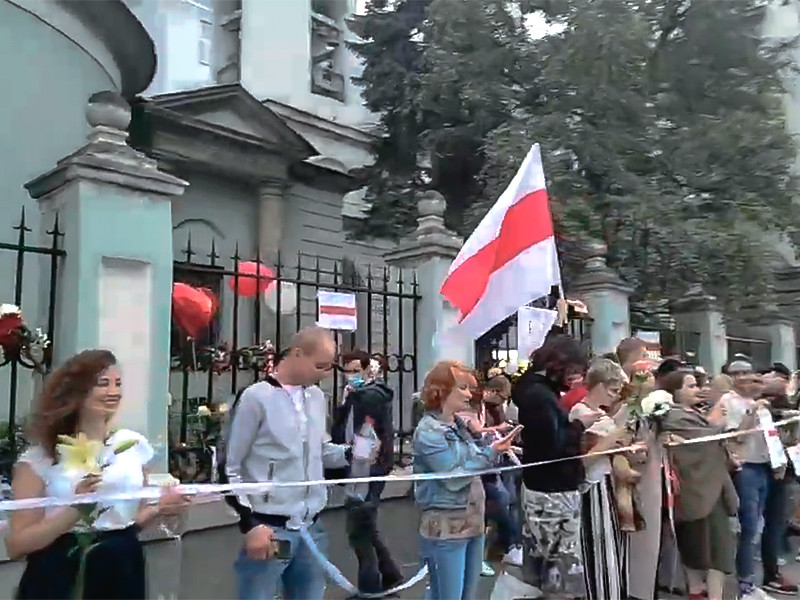 Активисты выстроились в цепочку солидарности напротив посольства на Маросейке. В руках собравшиеся держат белые и красные гвоздики и розы, плакаты с политическими лозунгами, воздушные шары
